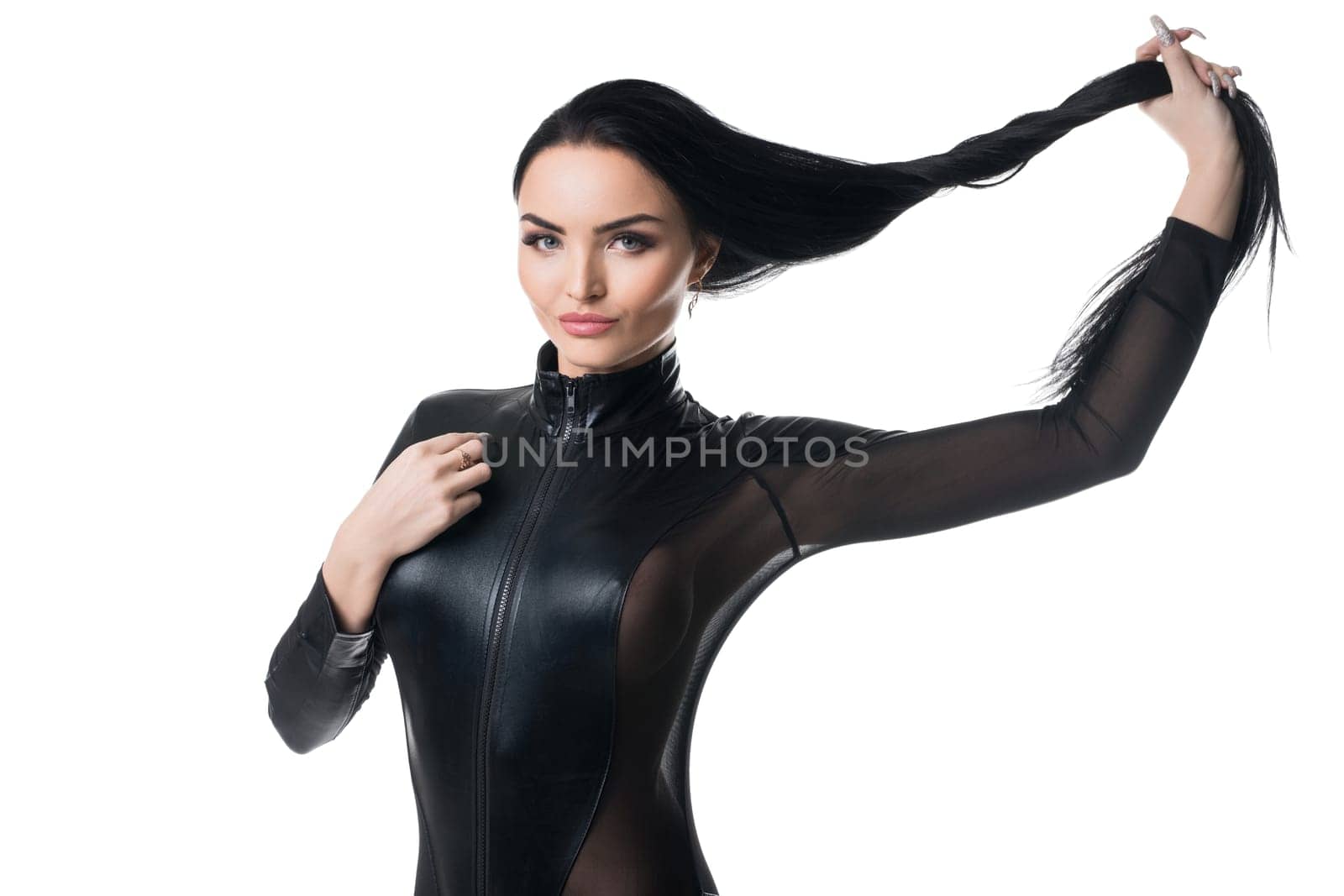 Seductive female in black leather bodysuit touching long hair and looking at camera on white background in studio