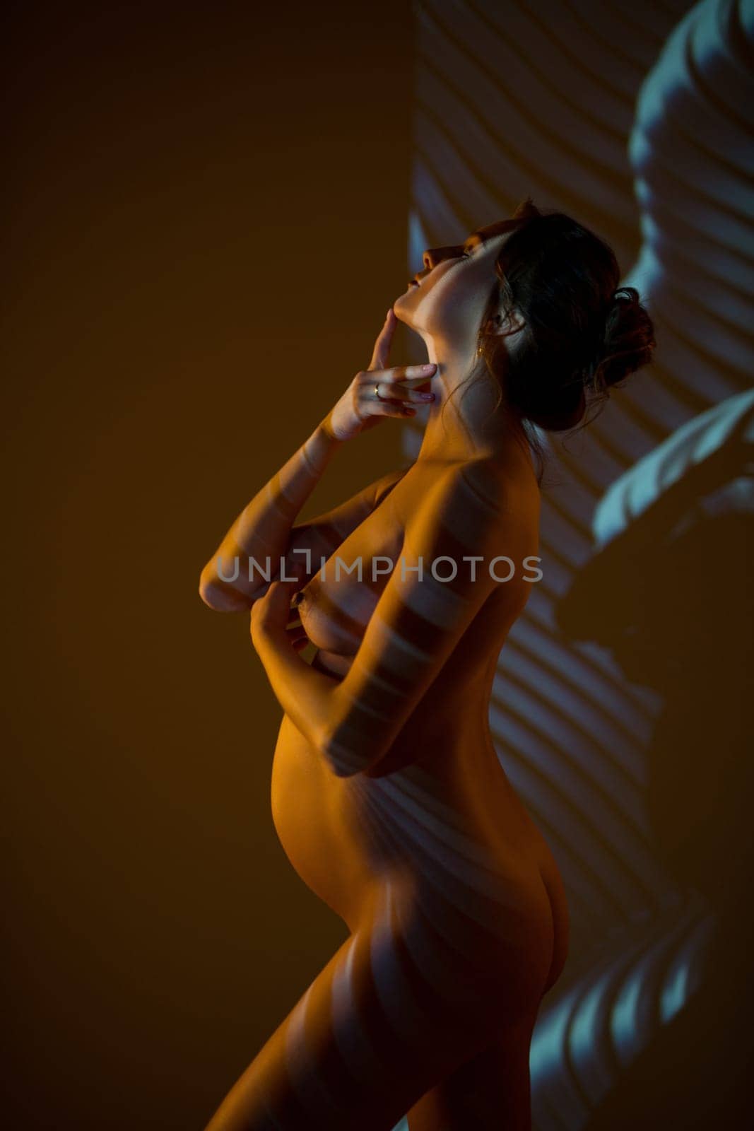 Side view of dreamy nude pregnant female touching chin while standing gracefully in dark studio with striped shadows and light