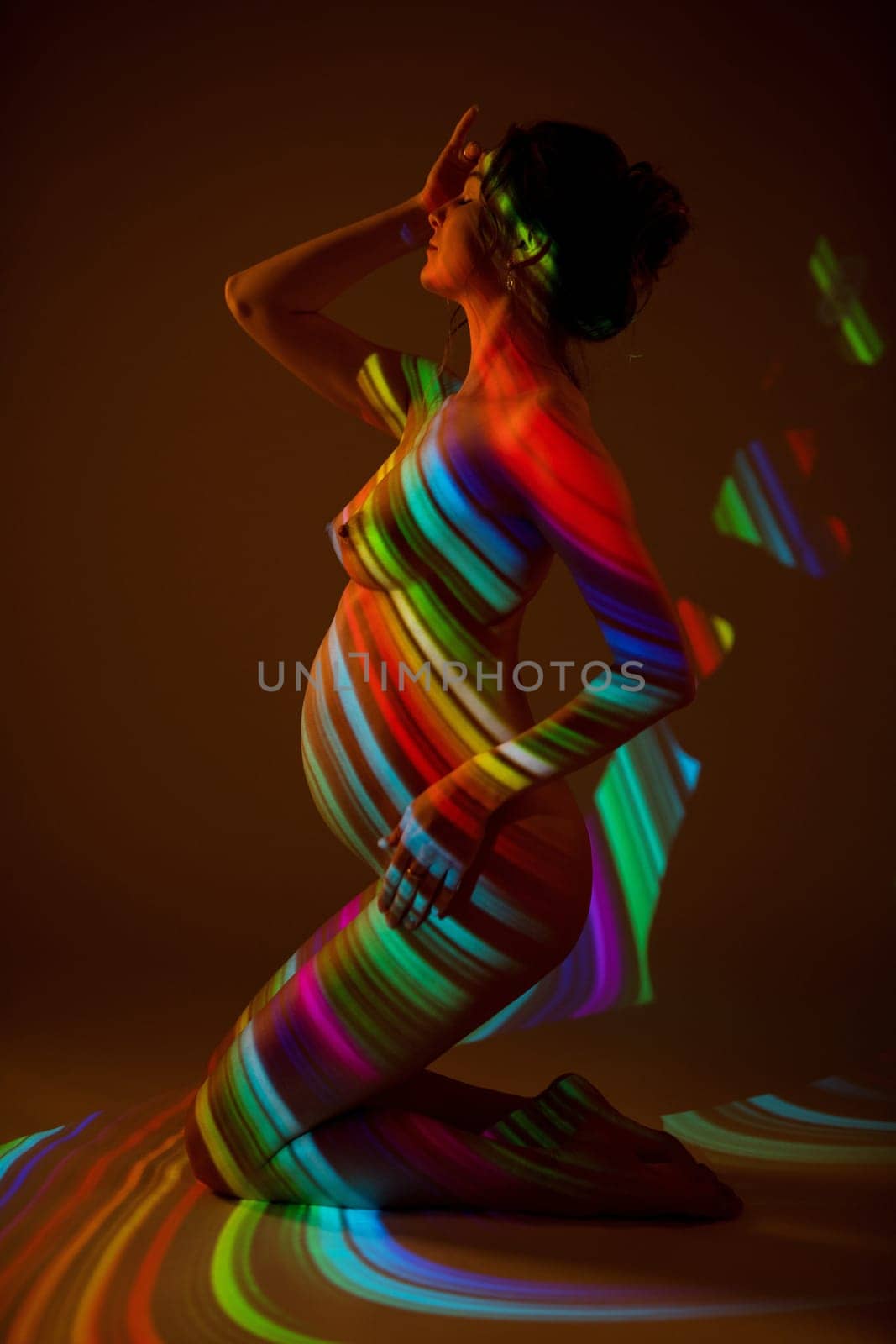 Side view of sensual nude pregnant female standing on knees in dark studio with colorful neon illumination