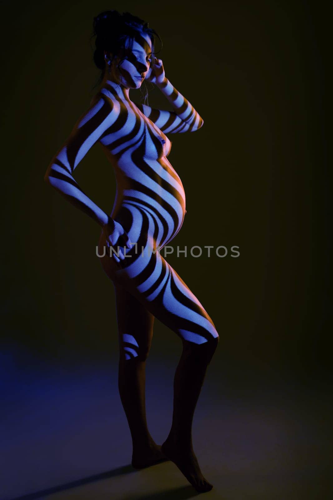 Side view of sensual nude pregnant female in dark studio with stripes shadows body paint