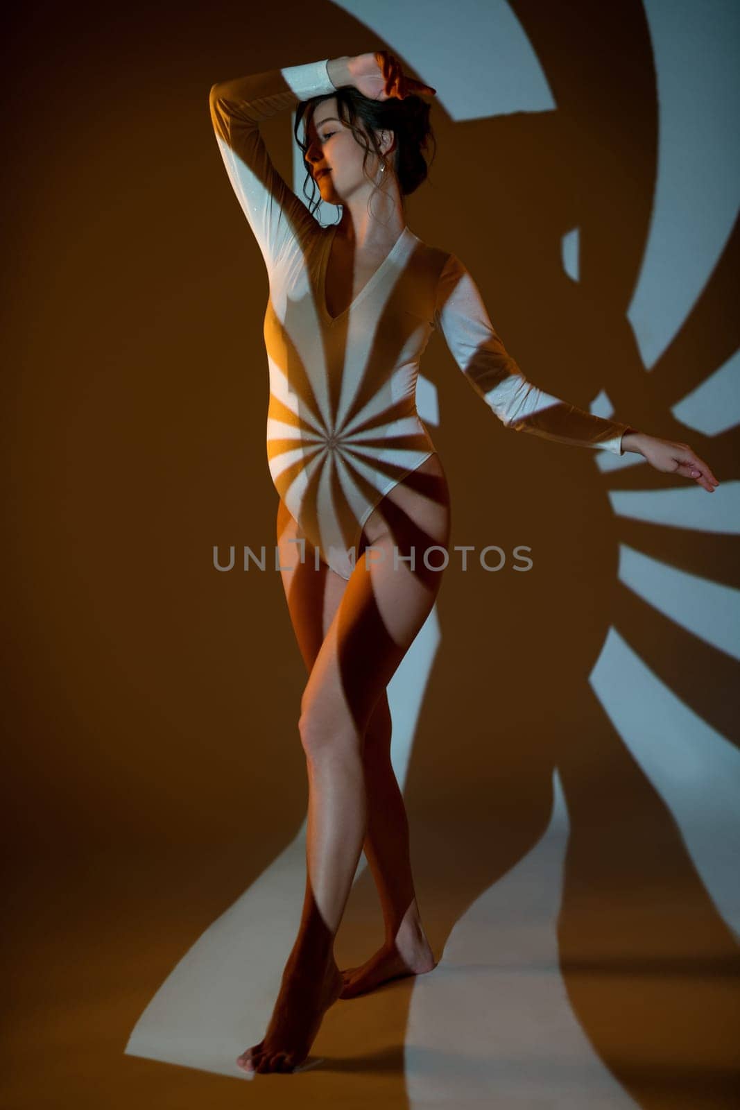 Graceful pregnant female in white bodysuit standing in dark studio illuminated by striped light of projector