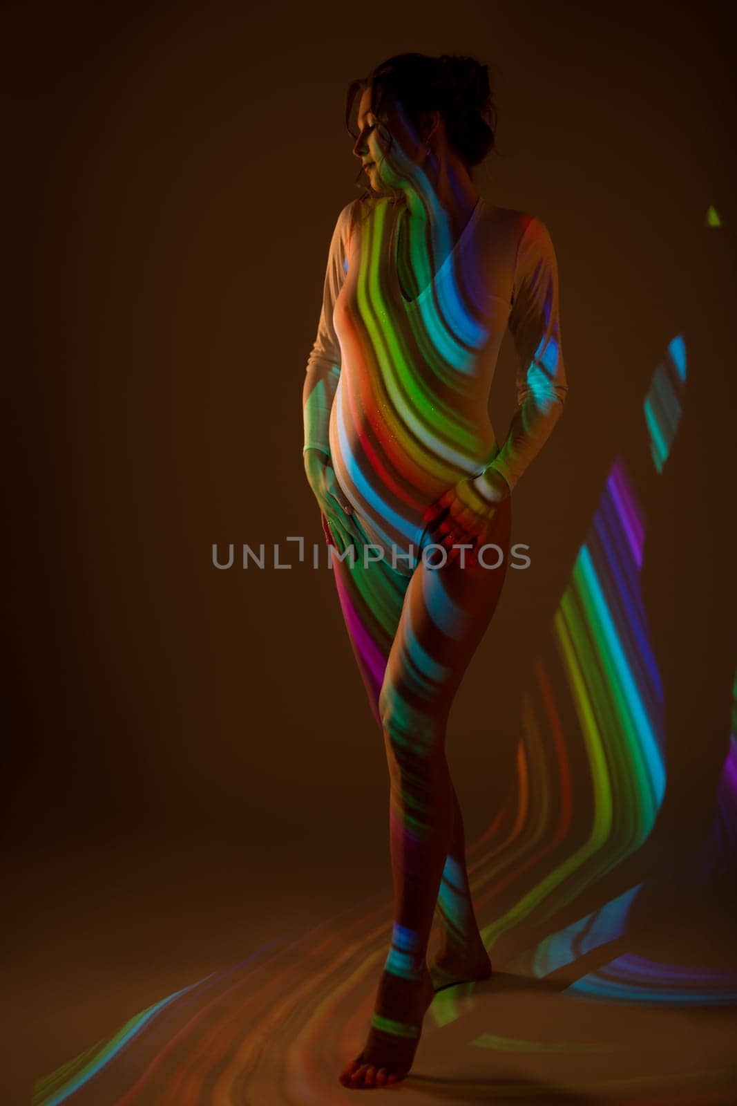 Graceful pregnant female in white bodysuit standing in dark studio illuminated by striped color light of projector