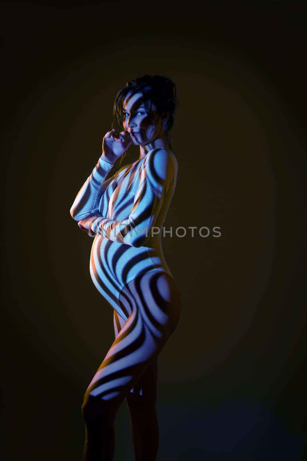 Side view of sensual nude pregnant female in dark studio with stripes shadows body paint