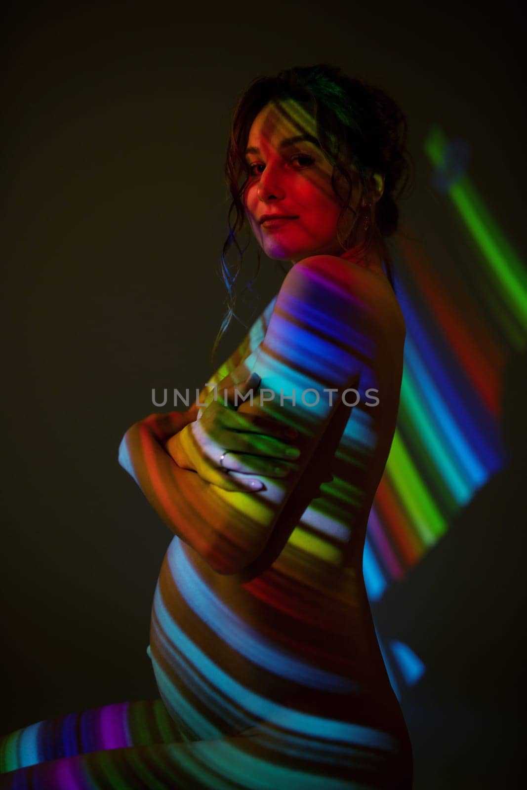 Side view of charming topless pregnant female covering breast with hands and looking at camera while standing under multicolored striped illumination in dark studio