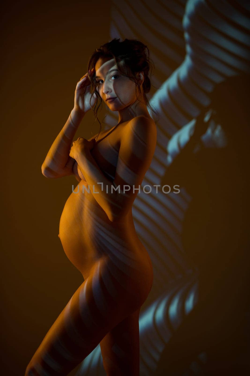 Side view of dreamy nude pregnant female touching chin while standing gracefully in dark studio with striped shadows and light