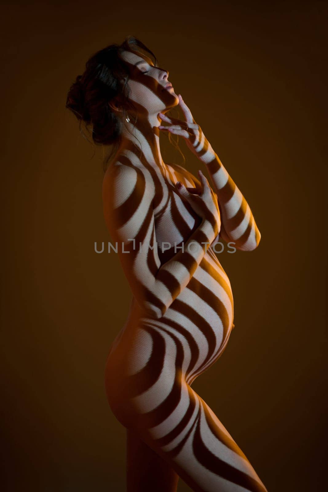 Side view of sensual nude pregnant female in dark studio with stripes shadows on body from projector