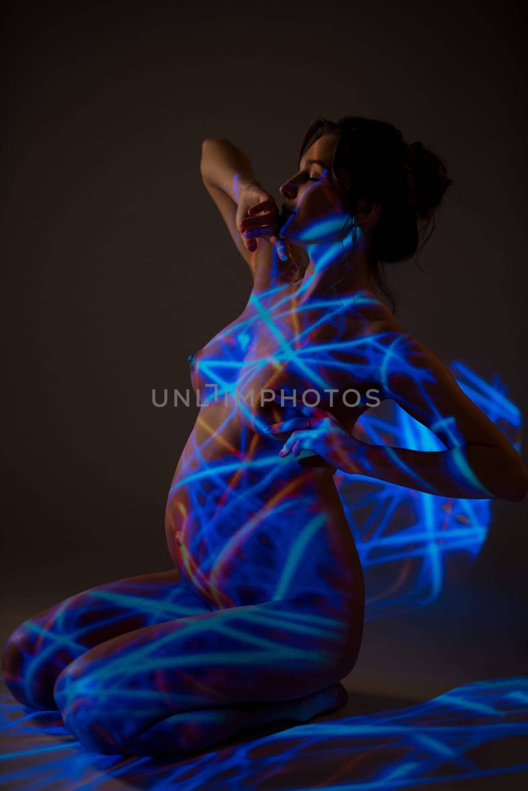Side view of naked pregnant female sitting with closed eyes on floor in dark studio with blue neon light