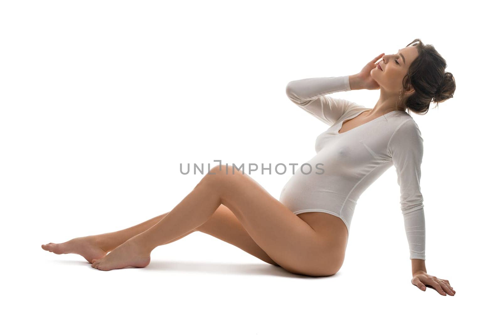 Full body side view of sensual gentle young pregnant female in white bodysuit kneeling and relax after stretching gracefully against white background