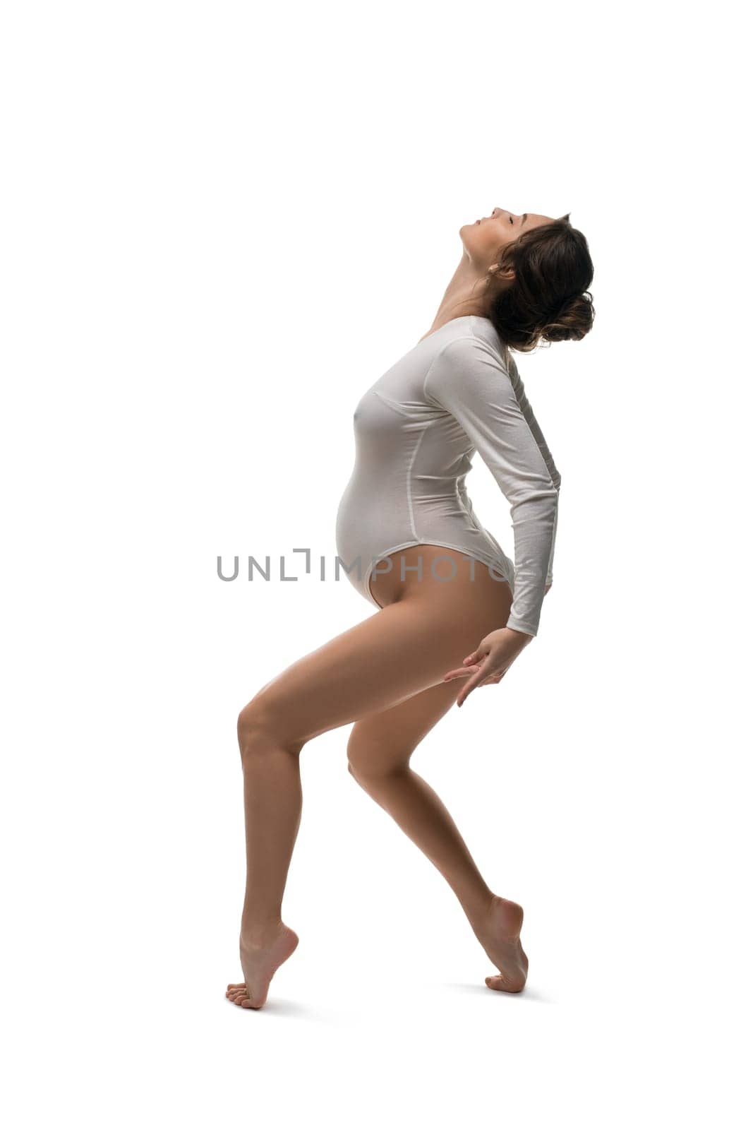 Side view of slim pregnant female in bodysuit standing on tiptoes on white background in studio