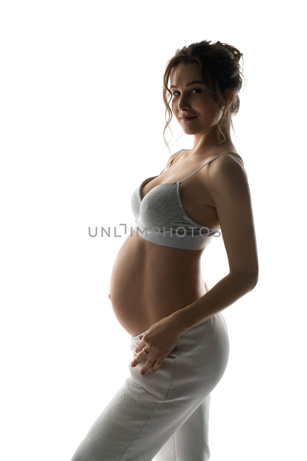 Side view of smiling young pregnant female in white bra and pants looking at camera while standing in light studio