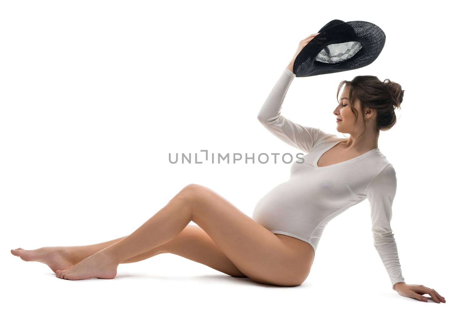 Full body side view of graceful young pregnant female in white bodysuit with black hat in hand sitting in white studio