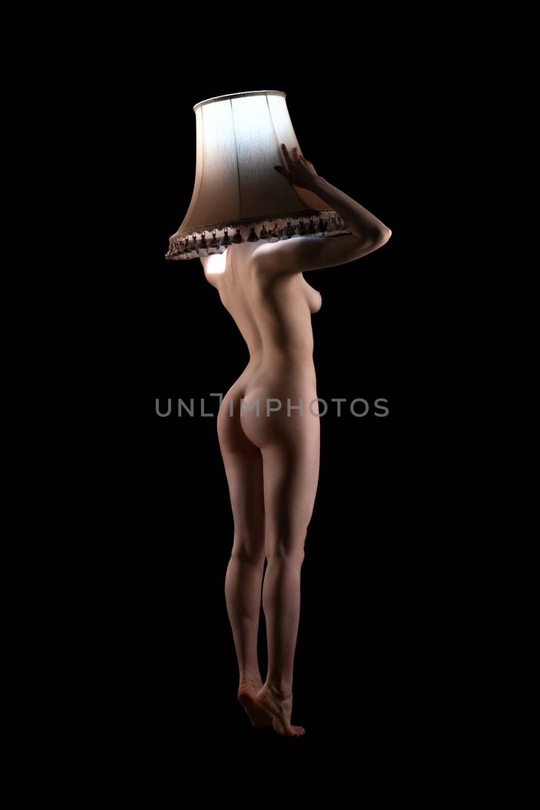 Full body of graceful nude slim female model holding lampshade over head - live lamp stand concept
