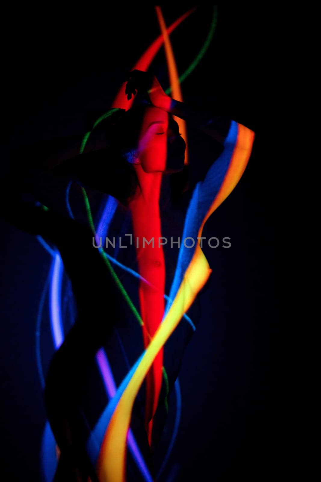 Artwork portrait of alluring young nude female model standing in dark studio with creative multicolored neon illumination