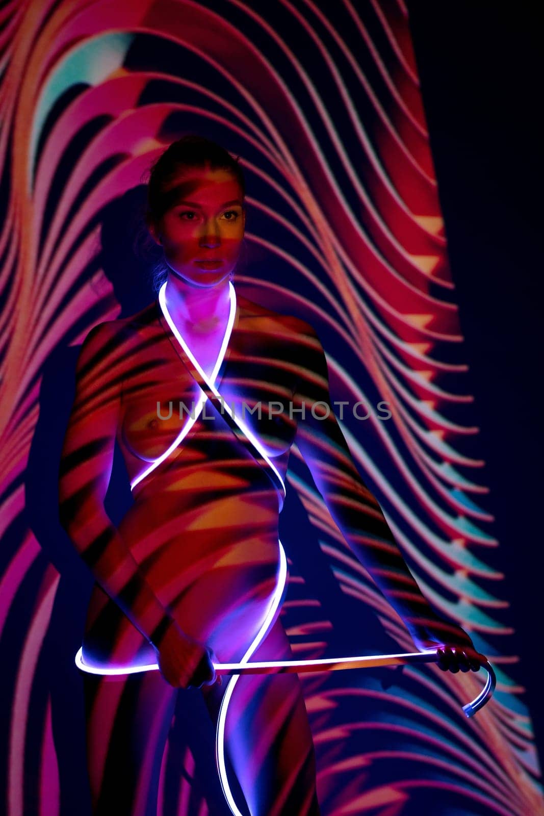 Artwork portrait of naked slim female model with bright neon strap standing under colorful projector illumination forming wavy pattern on body and wall