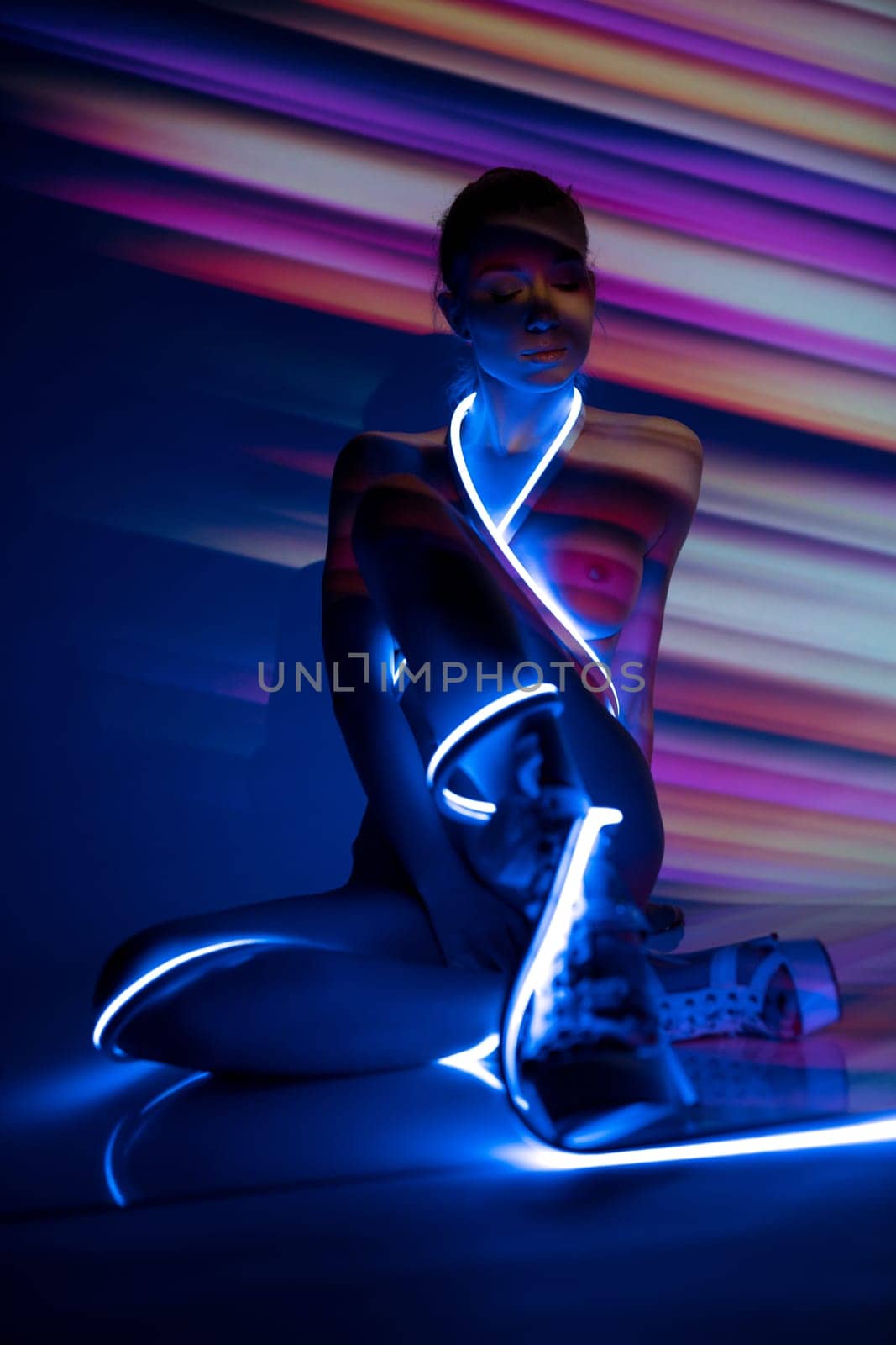 slim topless female model with glowing neon strap wrapped around body sitting under light of projector creating colorful striped pattern in studio