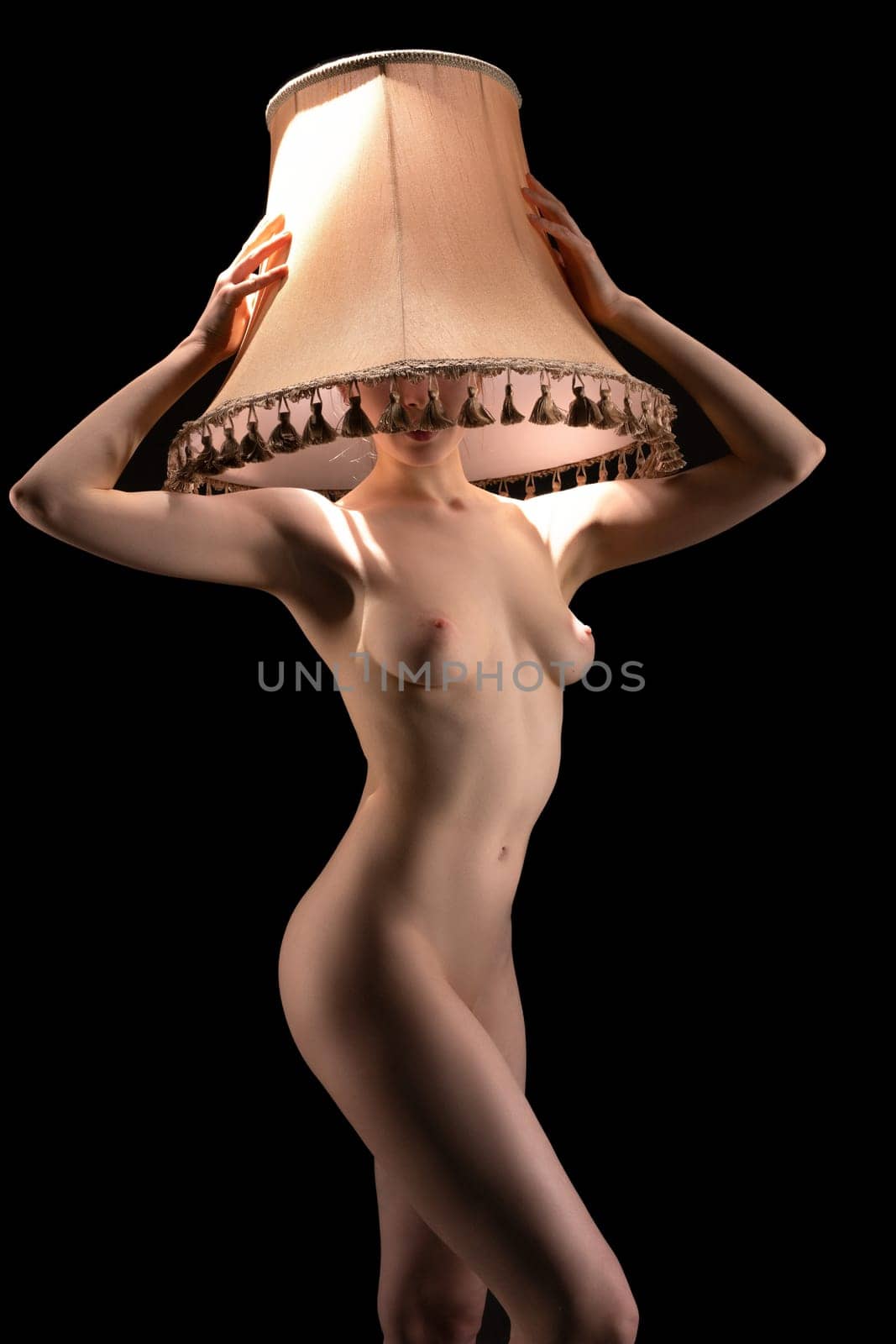 Slim curvy naked female model covering head and face with beige lampshade on black background