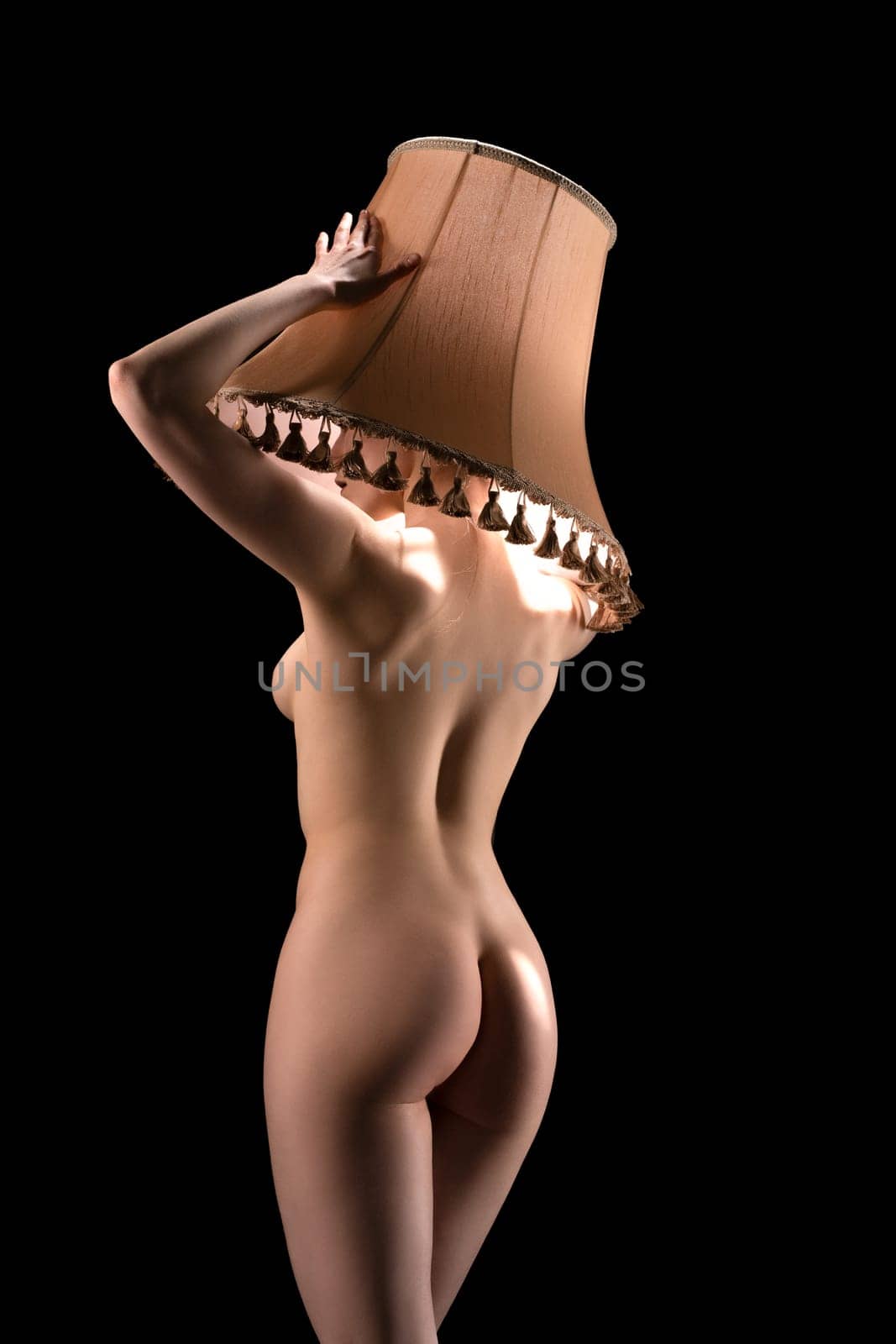Slim curvy naked female model covering head and face with beige lampshade back view