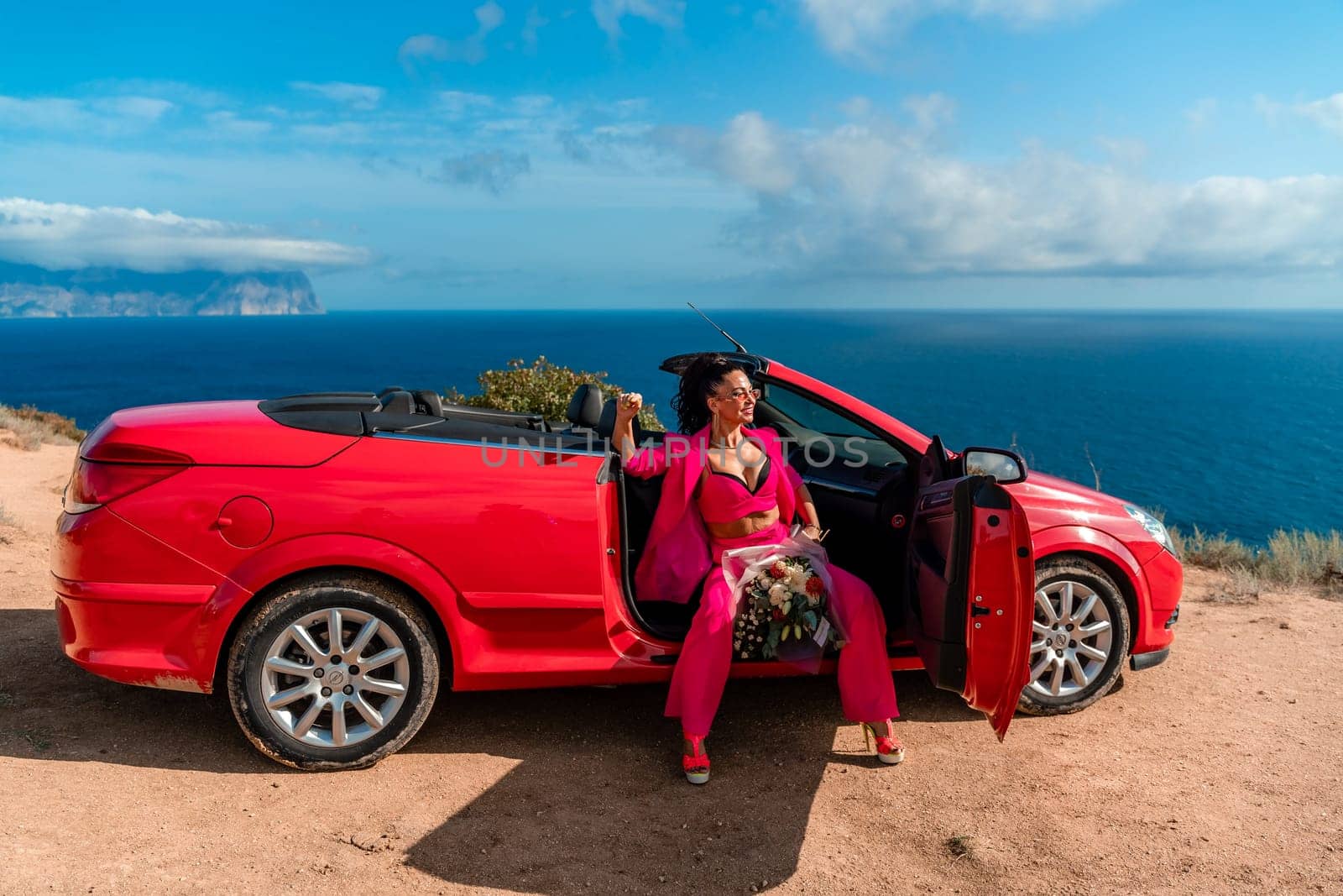 A woman is sitting in the back of a red convertible car. The car is parked on a dirt road near the ocean. The woman is wearing a pink suit and red shoes. Scene is relaxed and carefree