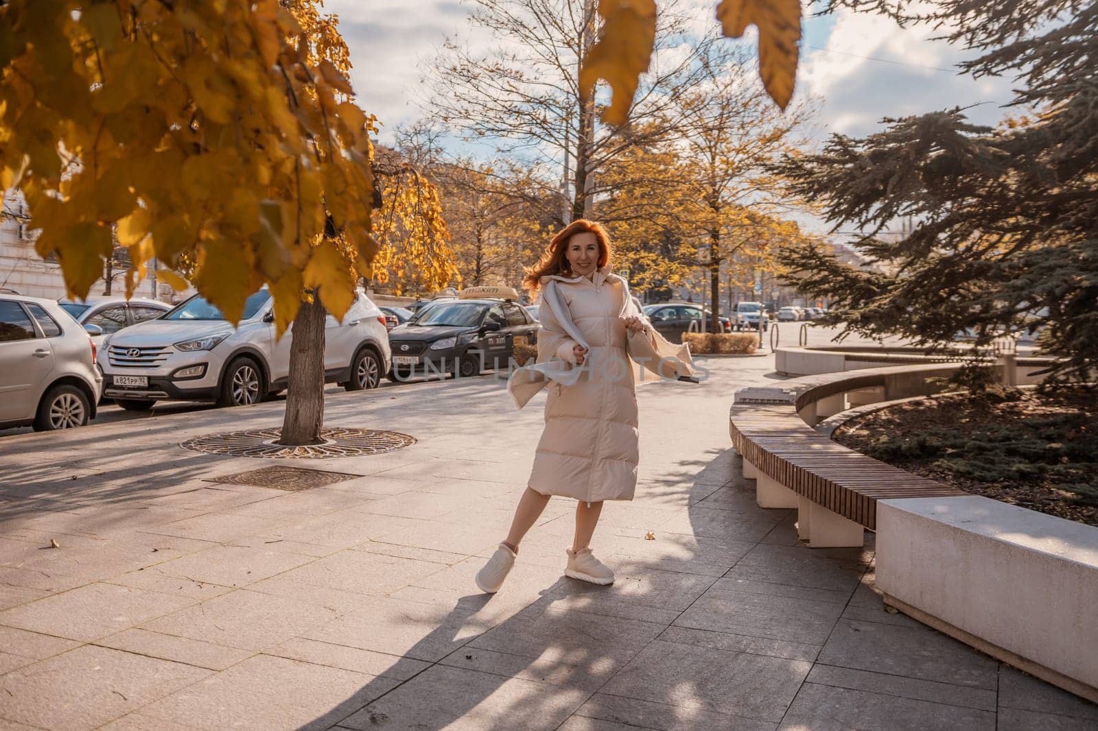 Woman in light dress playfully twirls on sunny city sidewalk, autumn leaves falling near parked cars. Bright sunlight casts shadows.