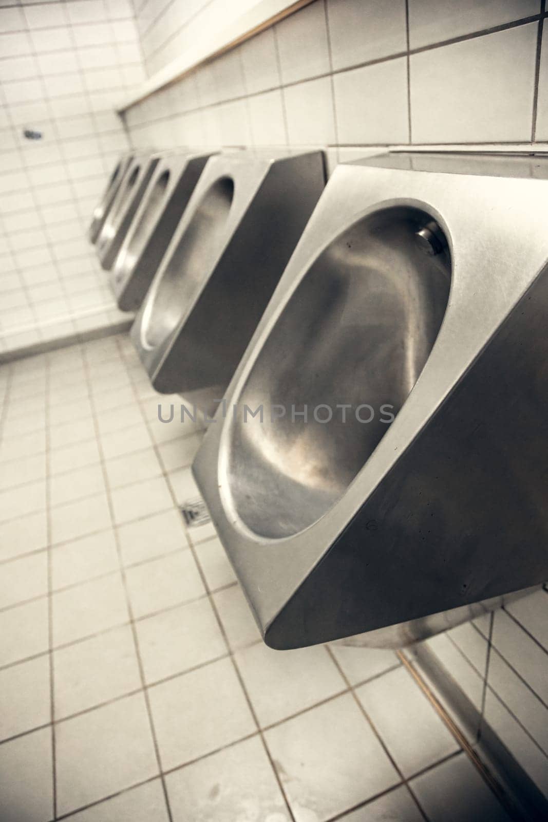 A row of stainless steel urinals in a public restroom with white tiled walls. The industrial, minimalist design highlights hygiene, sanitation, and modern restroom interiors