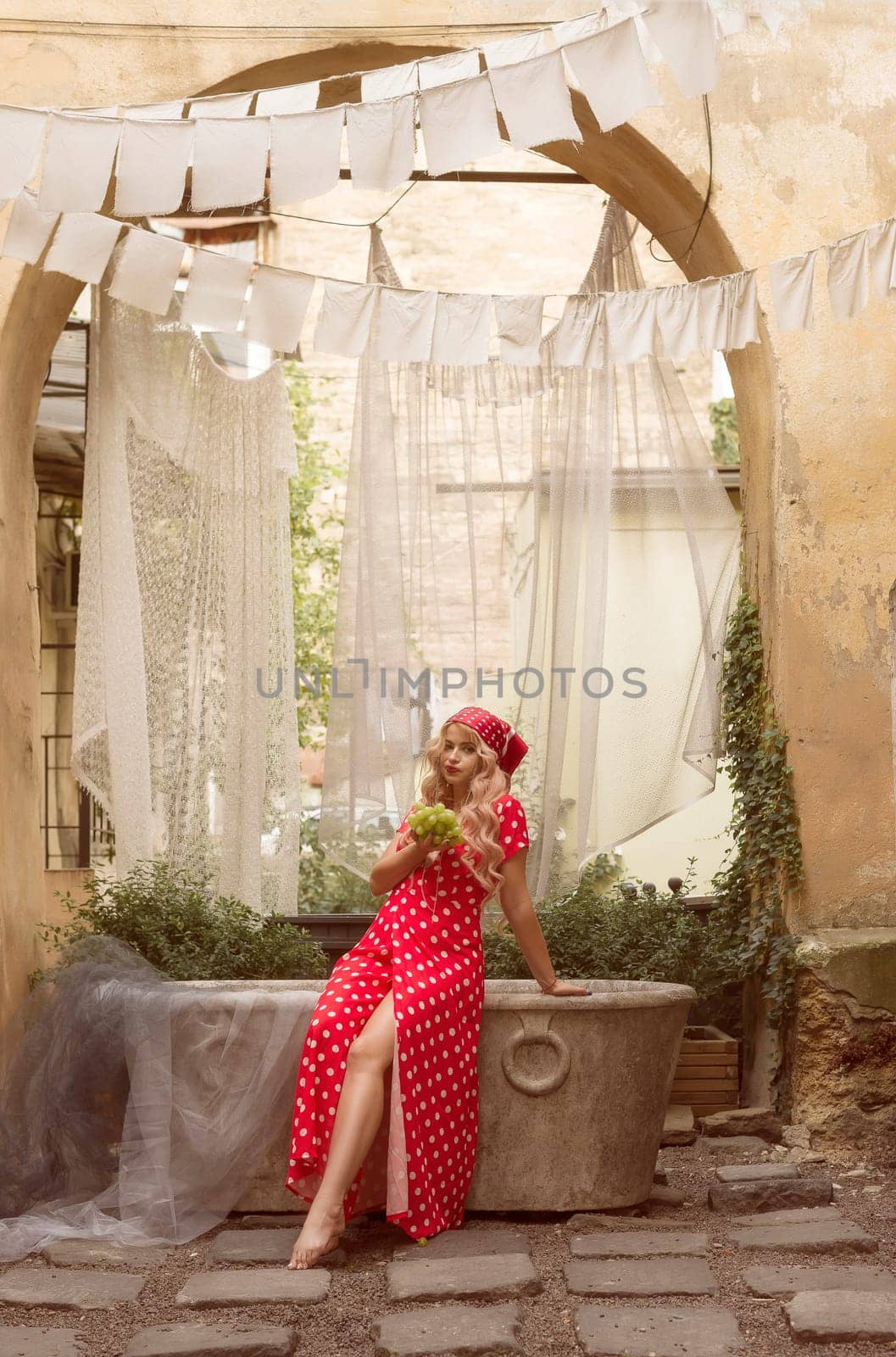 A young blonde woman is sitting in the courtyard of the old city, holding grapes in her hands. She is wearing a red dress with white polka dots and a headscarf.