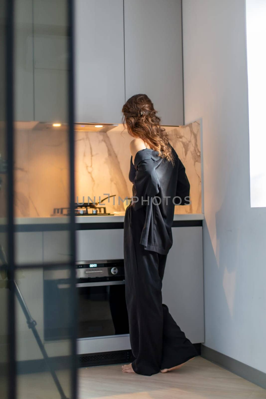 Woman in black silk pajamas cooks in a modern, minimalist kitchen with marble backsplash. Warm light illuminates the scene.