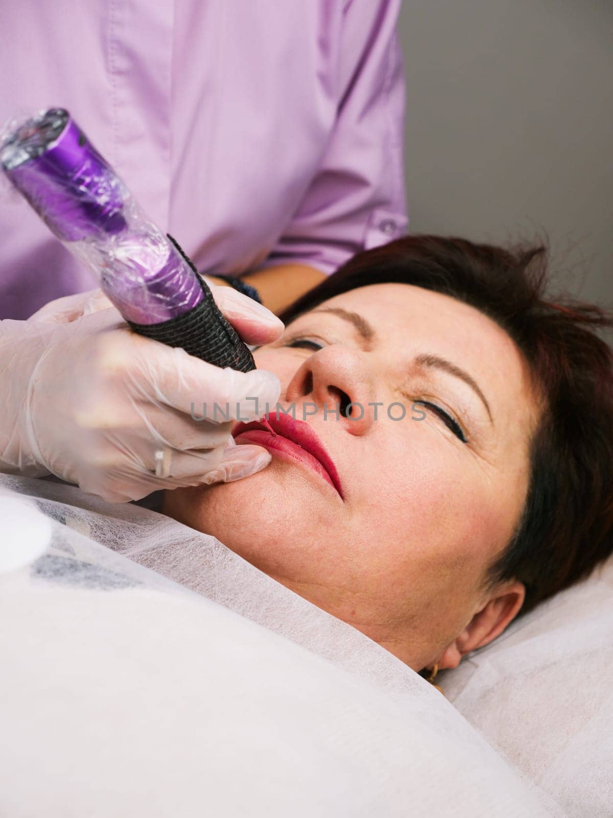 Permanent lip makeup beautiful woman over 50 years old in a beauty salon. A close-up beautician does a tattoo on his face. Vertical