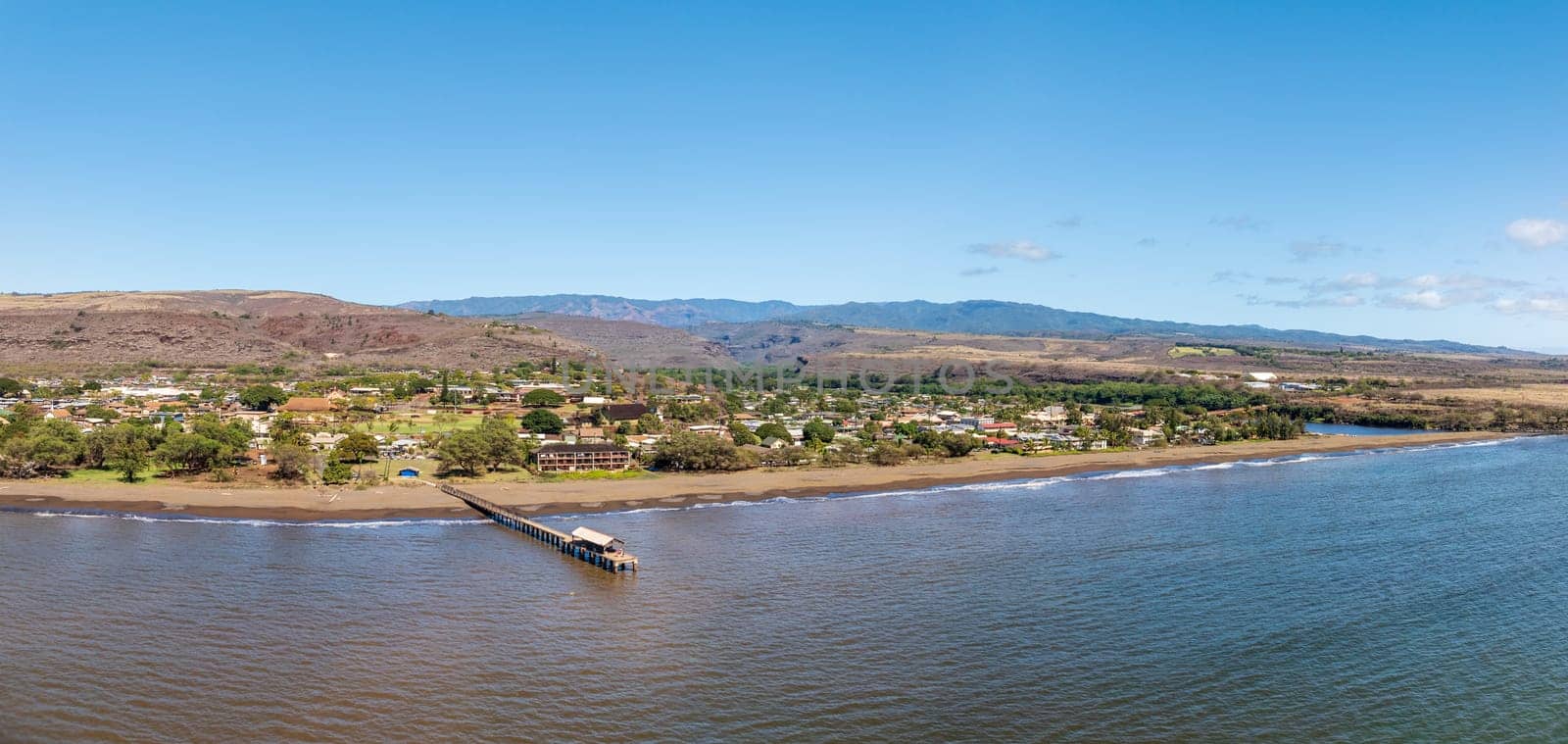 Panoramic aerial shot showing serene beauty of Waimea, Kauai showcasing its pier, coastline, and the landscape beyond under a clear sky