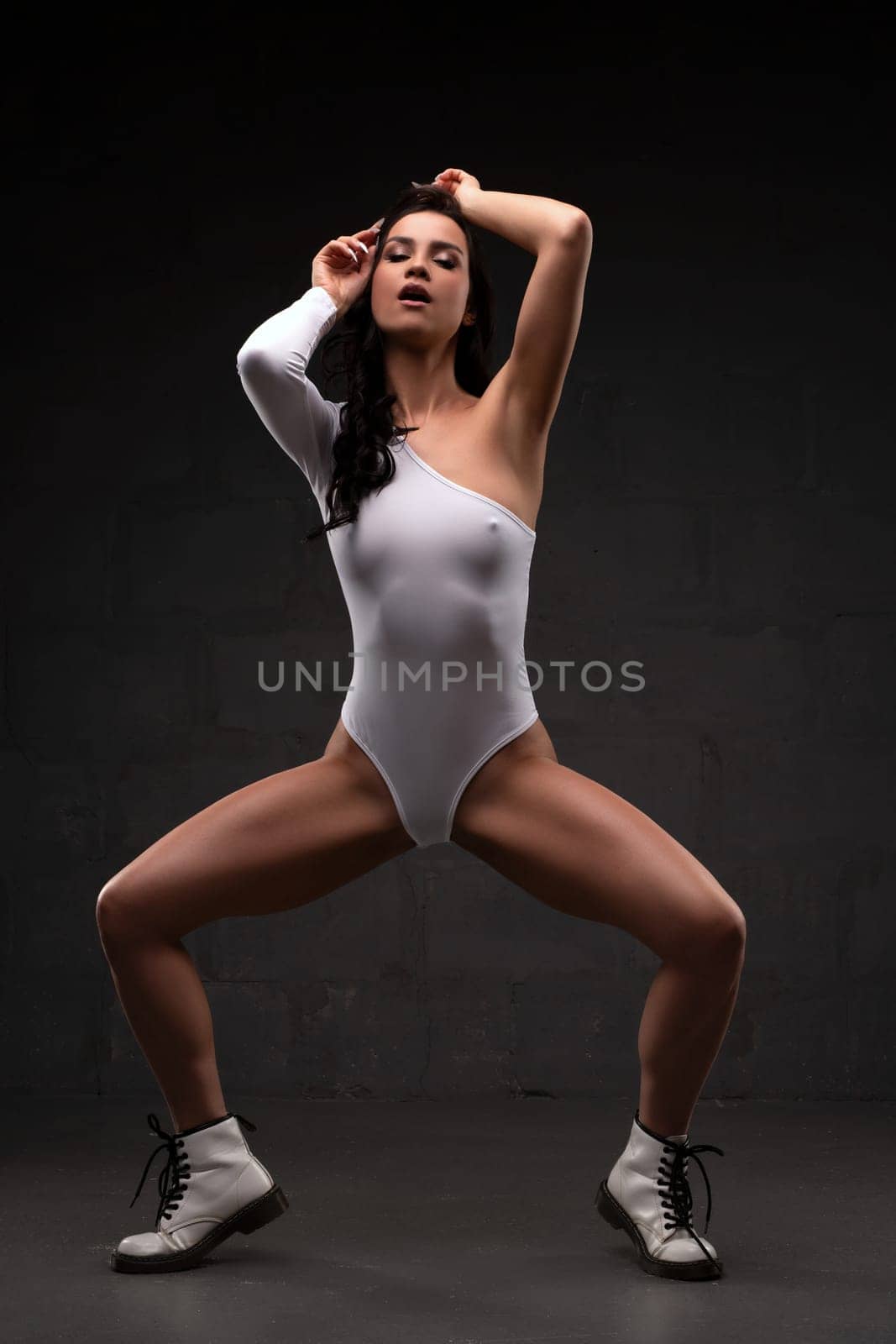 Full body slim young brunette in white bodysuit and leather boots standing in seductive provocative pose against gray background