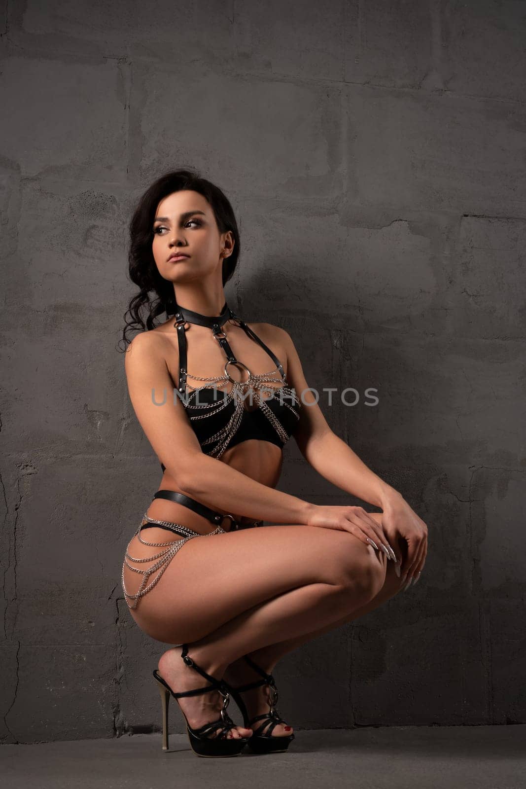 Full body of seductive young ethnic brunette in black leather lingerie with straps and chain and high heeled shoes sitting on floor against gray background