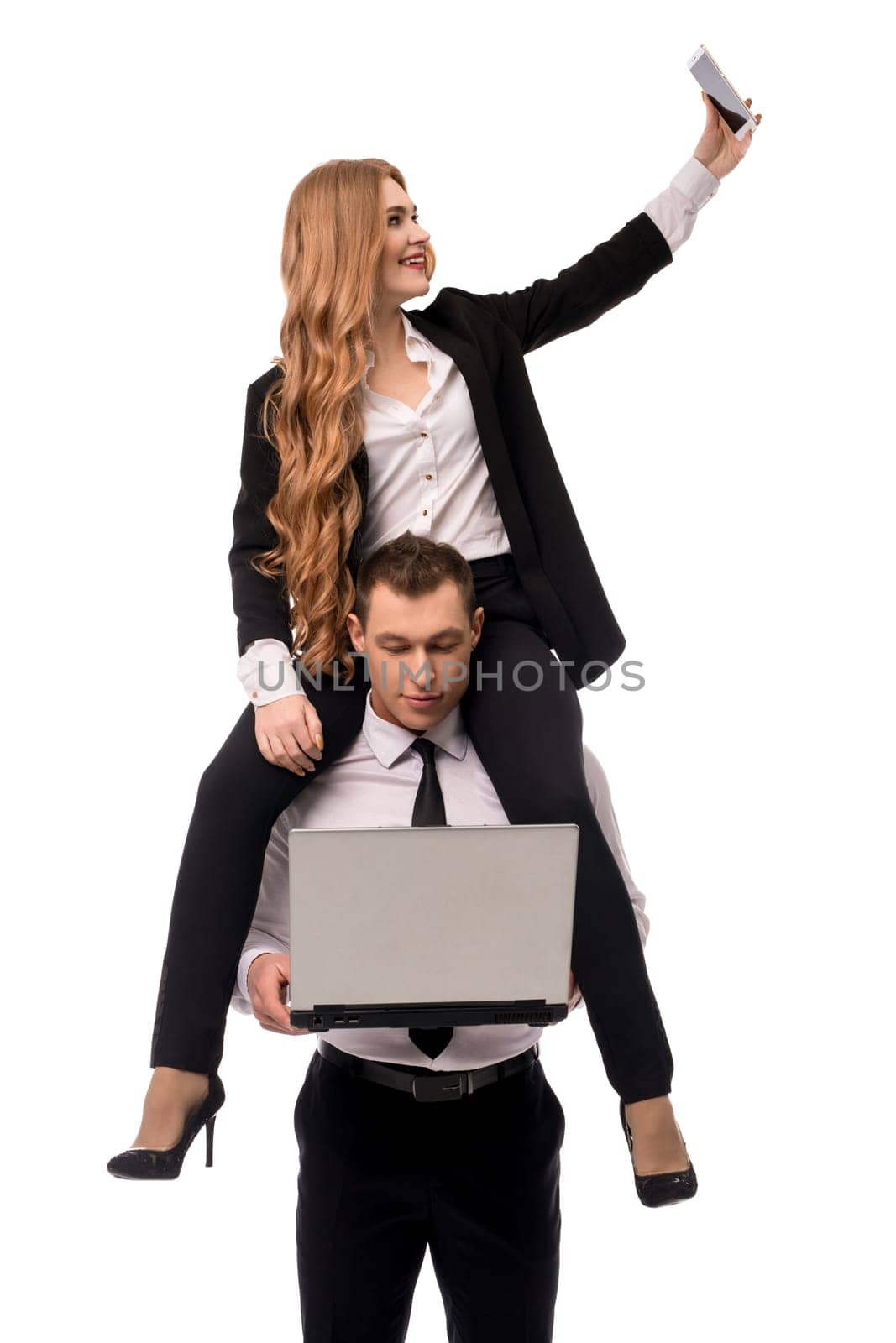 Businesswoman in formal wear doing selfie on smartphone while sitting on shoulders of businessman using laptop isolated on white background
