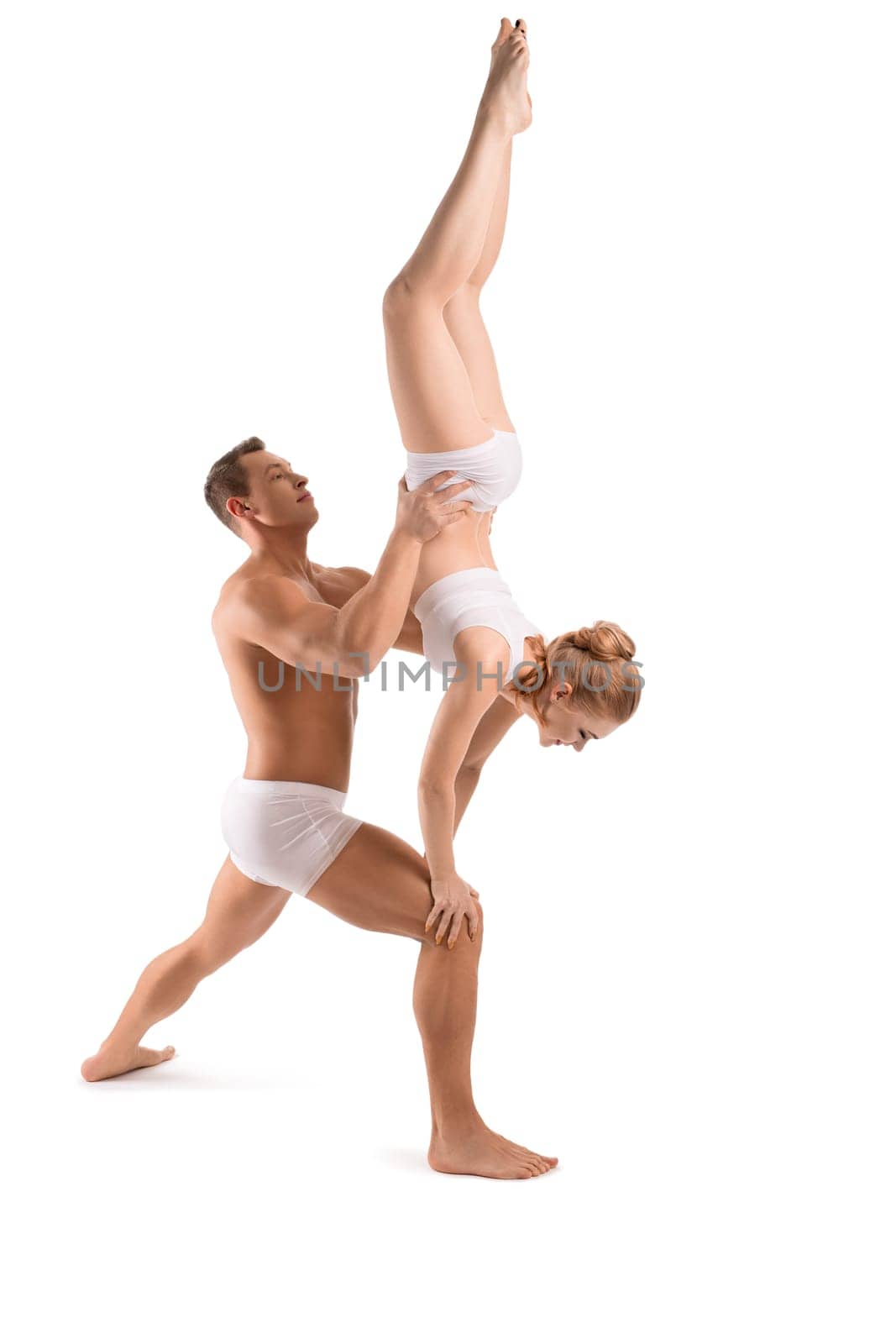 Full body side view of strong shirtless man standing in lunge position and supporting female partner doing handstand during acro yoga practice on white background