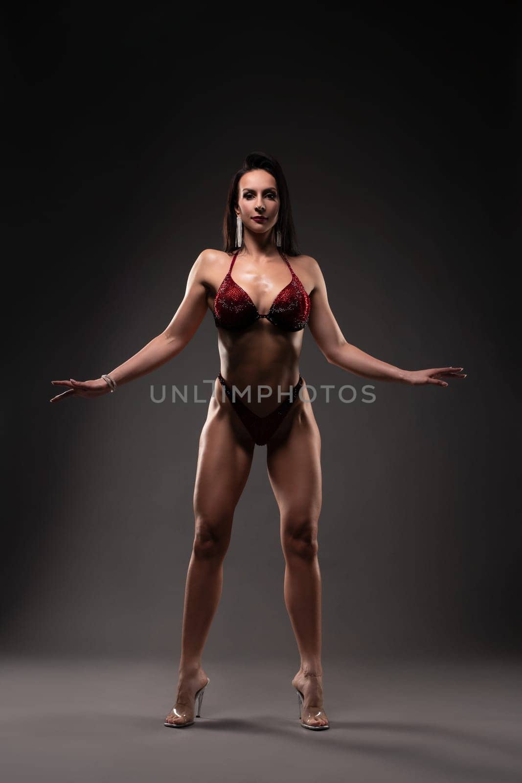 Strong brunette in red bra and panties ready for figure and fitness competition looking at camera against gray background