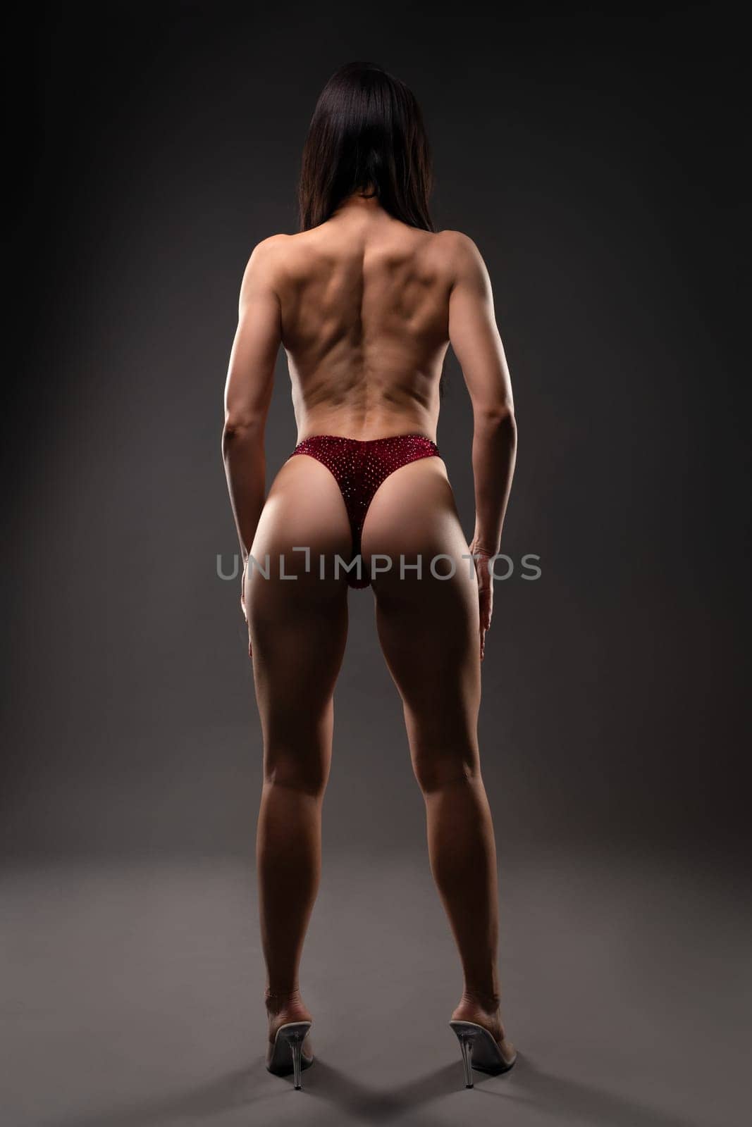 Full body of anonymous topless female with muscular back wearing thong and high heels against gray background
