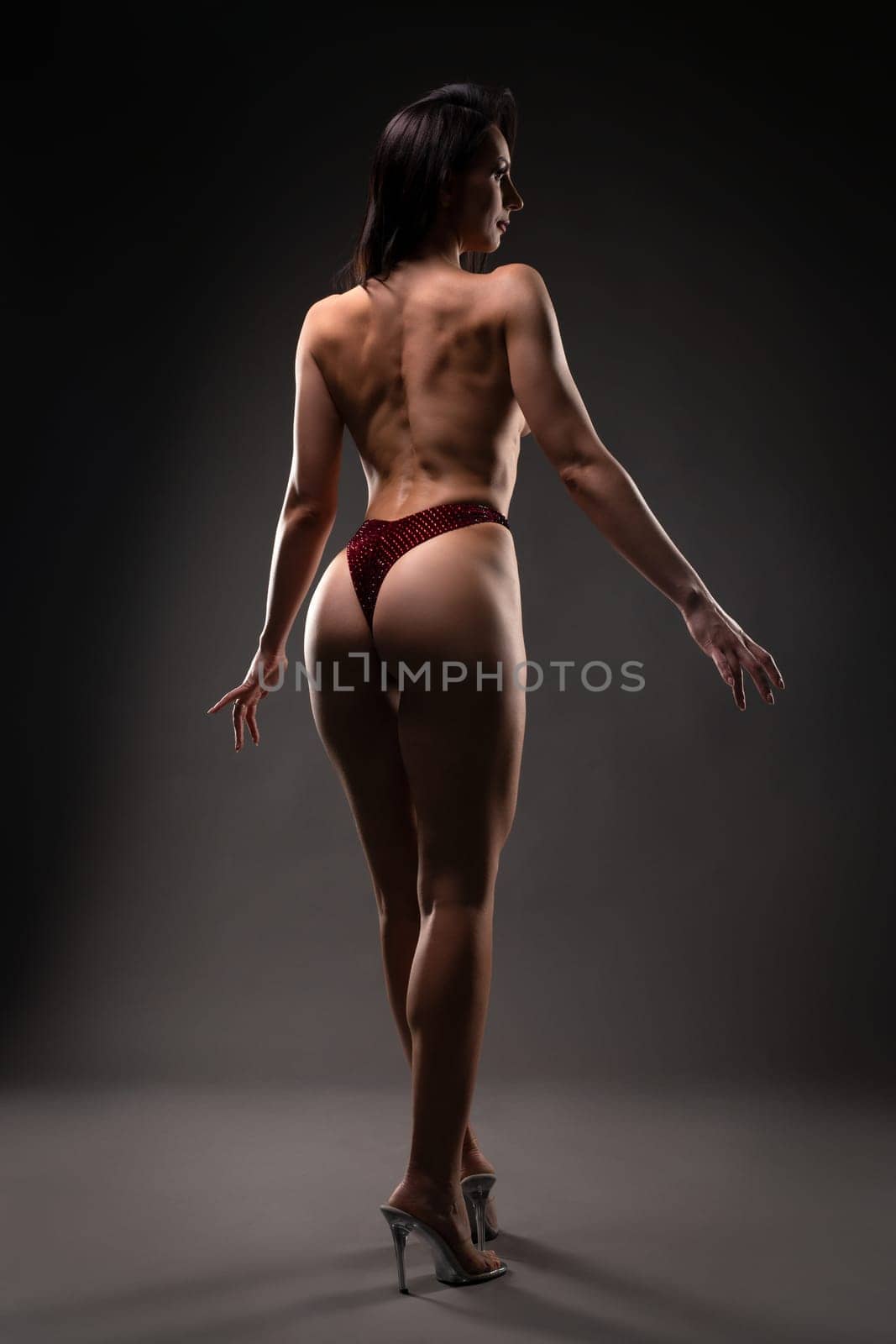 Full body of topless brunette female with muscular back wearing thong and high heels against gray background