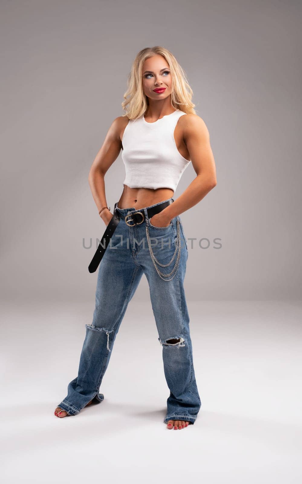 Muscular blond woman in jeans and white top looking at camera while stand on gray background