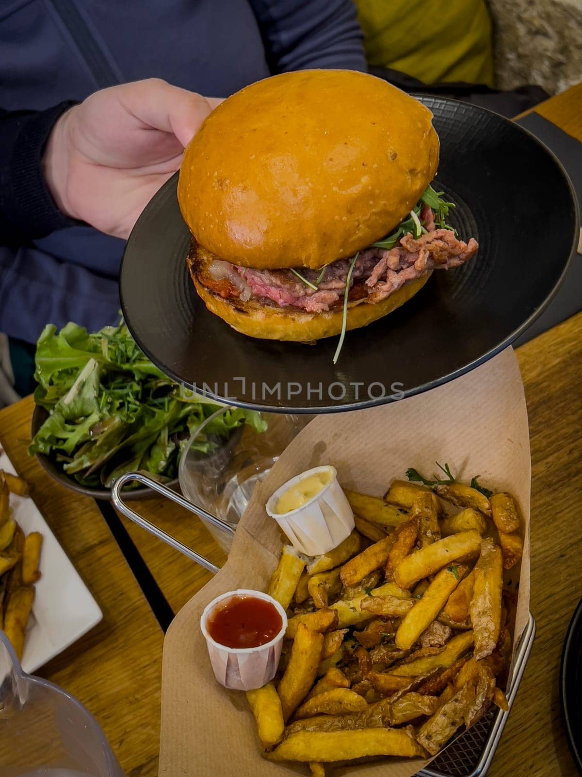 A person holds a freshly prepared burger topped with greens, accompanied by a generous serving of crispy fries and dipping sauces, creating a satisfying meal experience.