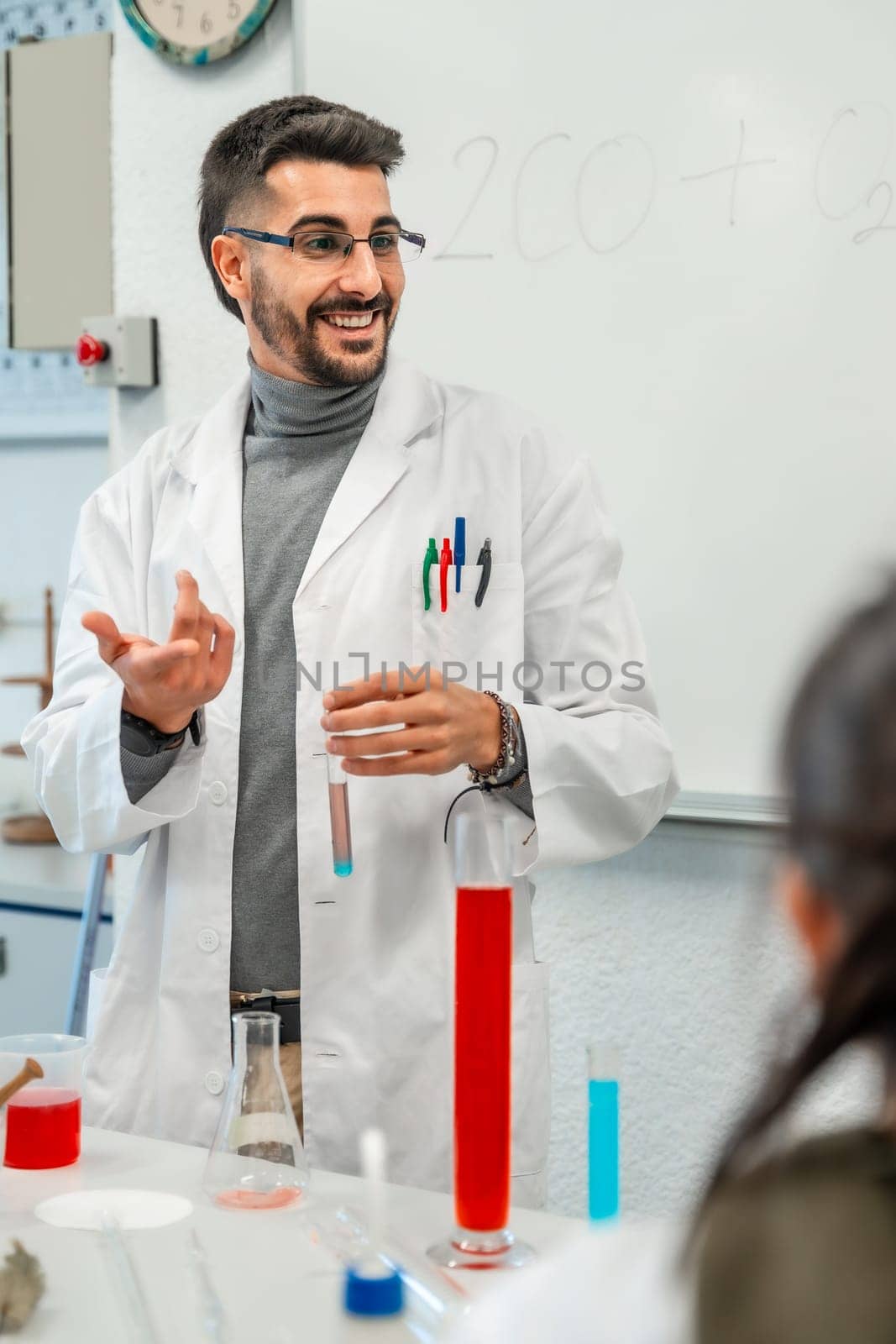 Smiling chemistry teacher showing colorful test tubes and beakers while explaining chemical experiment to students in a university laboratory