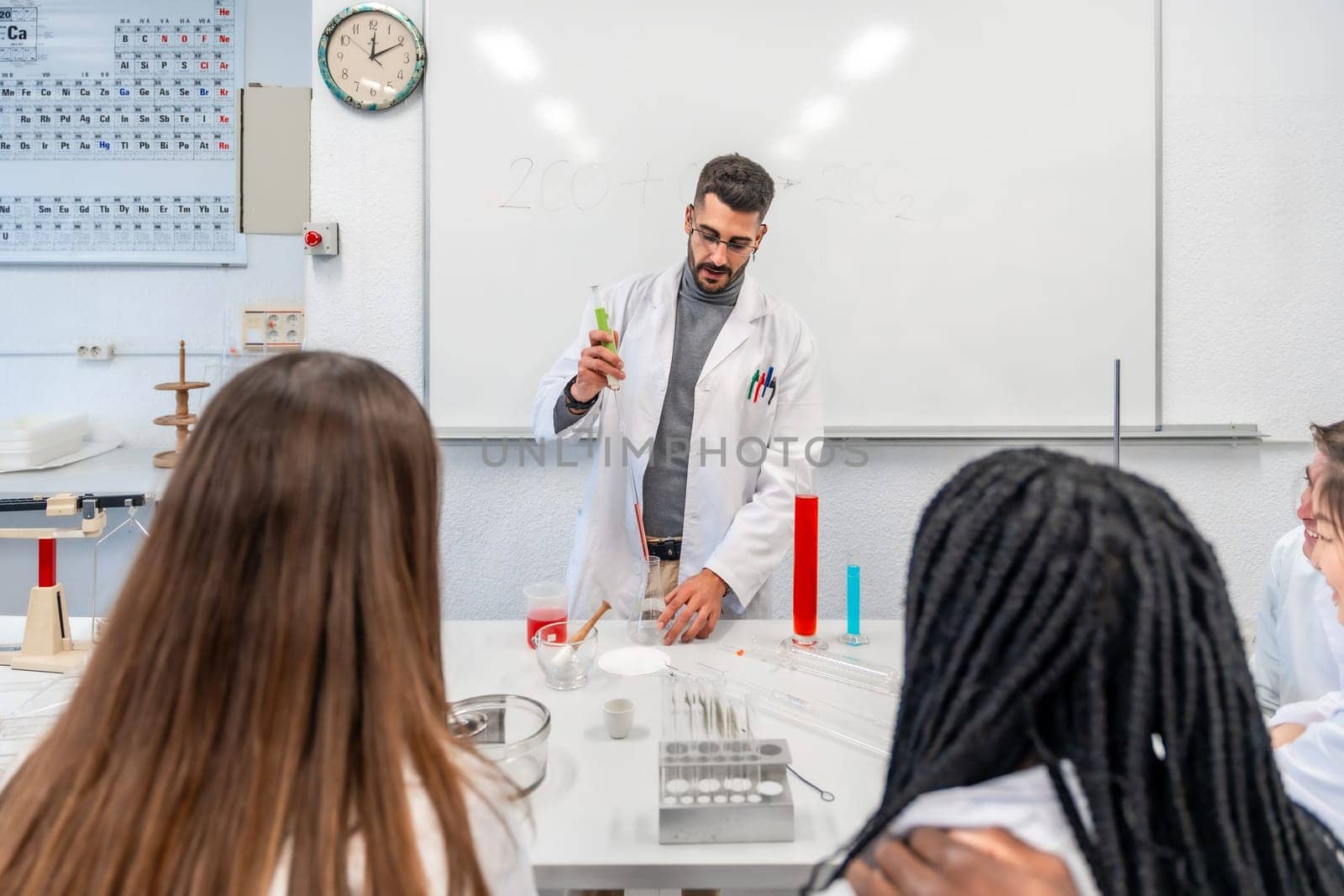 University teacher wearing lab coat performing a chemistry experiment with colorful liquids in beakers and test tubes, in front of multi ethnic students
