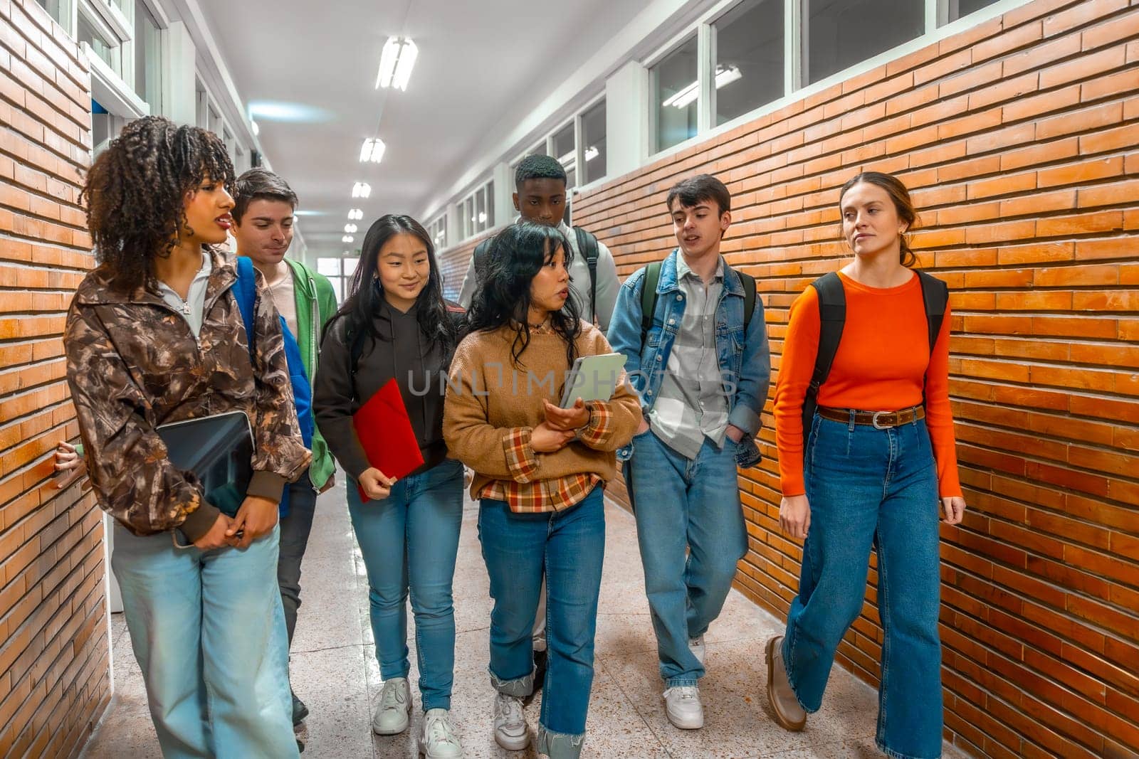 Multi ethnic university students walking down school hallway, chatting and holding books and tablets
