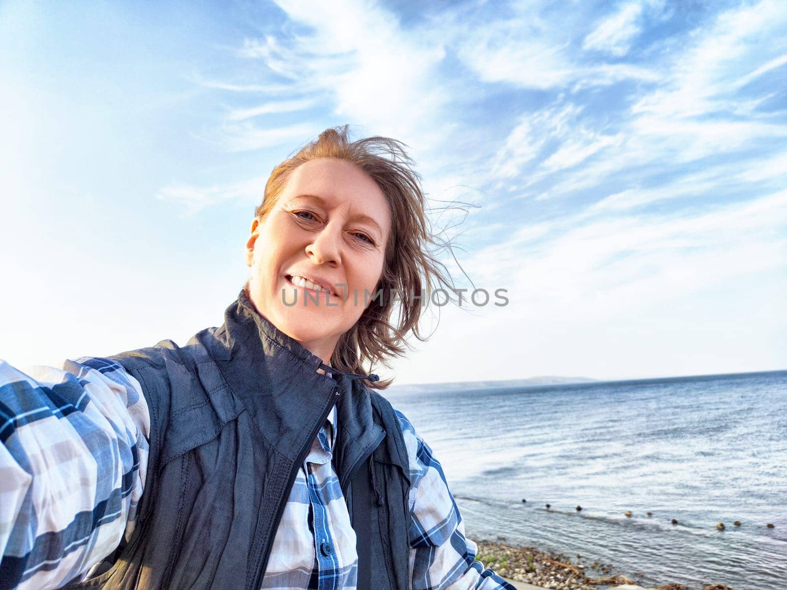 A middle-aged travel blogger snaps a cheerful seaside selfie on a sunny day.
