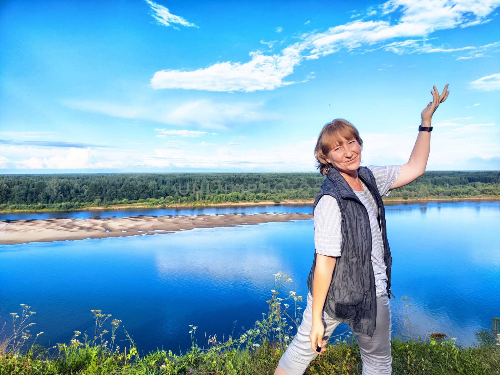 A blonde middle-aged woman takes a selfie smiling by a beautiful river landscape during a summer trip to nature.