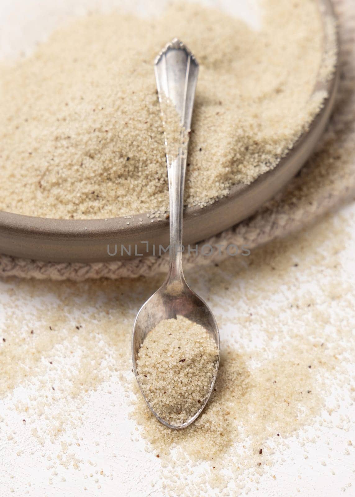 Raw uncooked fonio seeds in a plate with a metal spoon on white table close up, copy space. African ancient pseudocereal. Organic super food. Healthy food concept. Gluten free alternative seeds