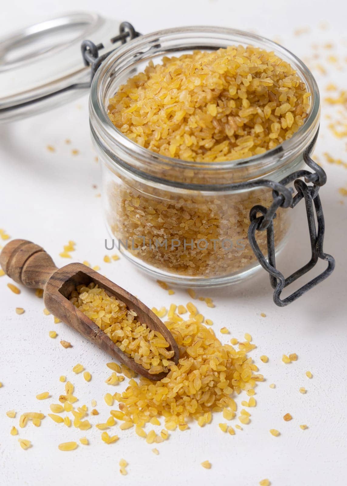 Opene glass jar of raw dry uncooked yellow bulgur grain with a wooden scoop on napkin closeup. Middle Eastern cousine. Organic super food for healthy diet. Ancient alternative cereal seeds