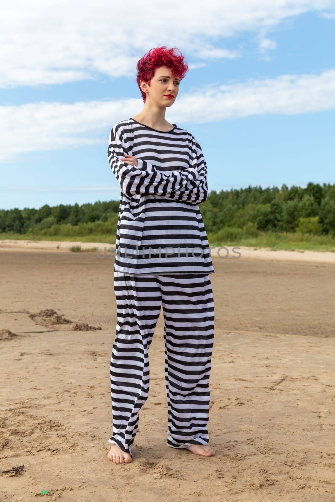 A young adult with curly red hair poses on a sandy beach, dressed in a loose striped outfit. The bright sky and surrounding trees add to the natural beauty of the setting.