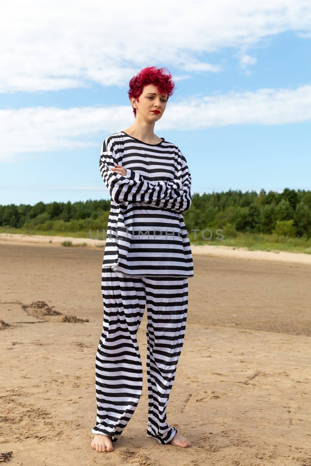 A person with vibrant red hair strikes a confident pose on a sandy beach. They wear a comfortable striped outfit, standing barefoot under a clear blue sky amidst lush green trees.