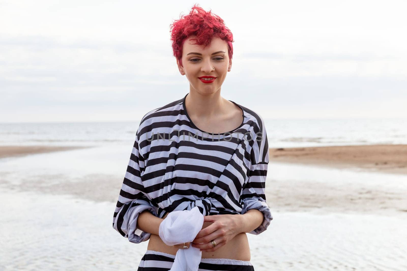 A woman stands on the beach with pink hair and a striped top, holding a towel. The shoreline features wet sand and gentle waves, with a cloudy sky in the background.