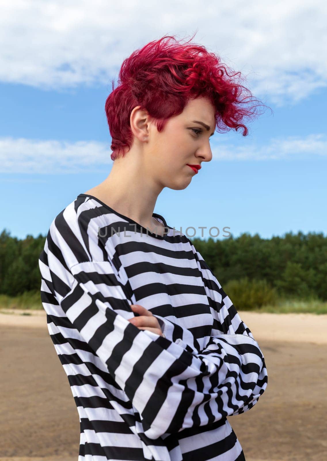 Individual with vibrant pink hair and a striped outfit stands outdoors, gazing thoughtfully at the landscape. The setting features a clear sky and lush greenery in the background.