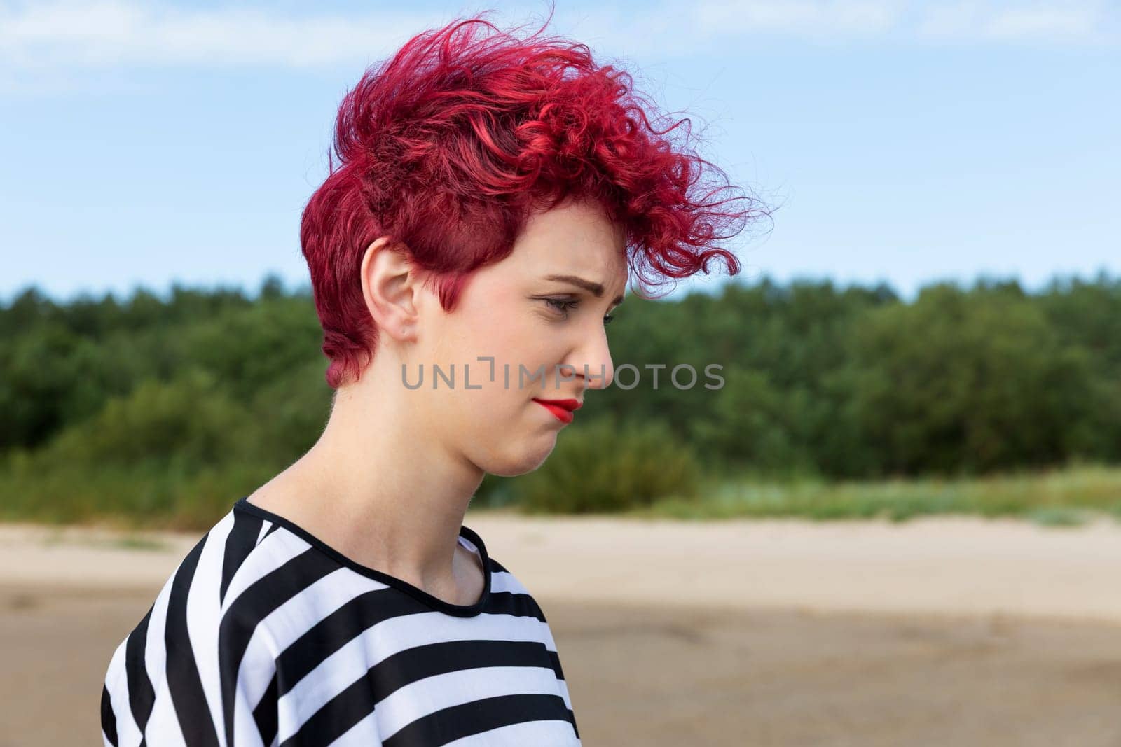 A young woman with vibrant red, curly hair strolls along a sandy beach. She wears a black and white striped top, reflecting a casual beach atmosphere on a clear day.