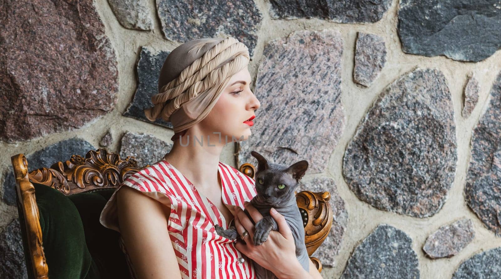 A woman dressed in vintage clothing sits gracefully with a cat in her arms. She gazes thoughtfully to the side, with a stylish hat adorning her head.